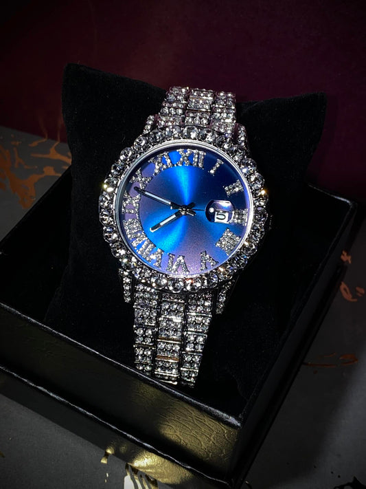 Silver-tone women's watch with a diamond-studded bezel and Roman numeral markers on a blue dial. The watch features a date display at the 3 o'clock position and a sparkling, crystal-encrusted band for a sophisticated, luxurious look.