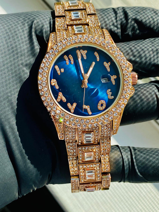 Gold-tone women's watch with diamond-studded bezel, featuring a blue dial with unique, stylized numbers and a date display. The watch has a sparkling, crystal-encrusted band for a glamorous, luxurious look.