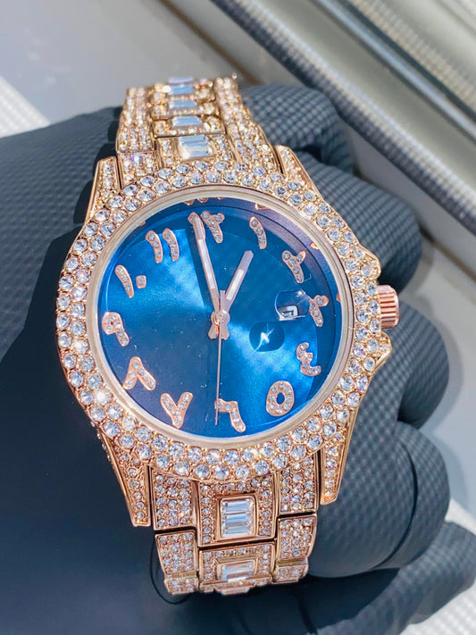 Gold-tone women's watch with diamond-studded bezel, featuring a blue dial with unique, stylized numbers and a date display. The watch has a sparkling, crystal-encrusted band for a glamorous, luxurious look.