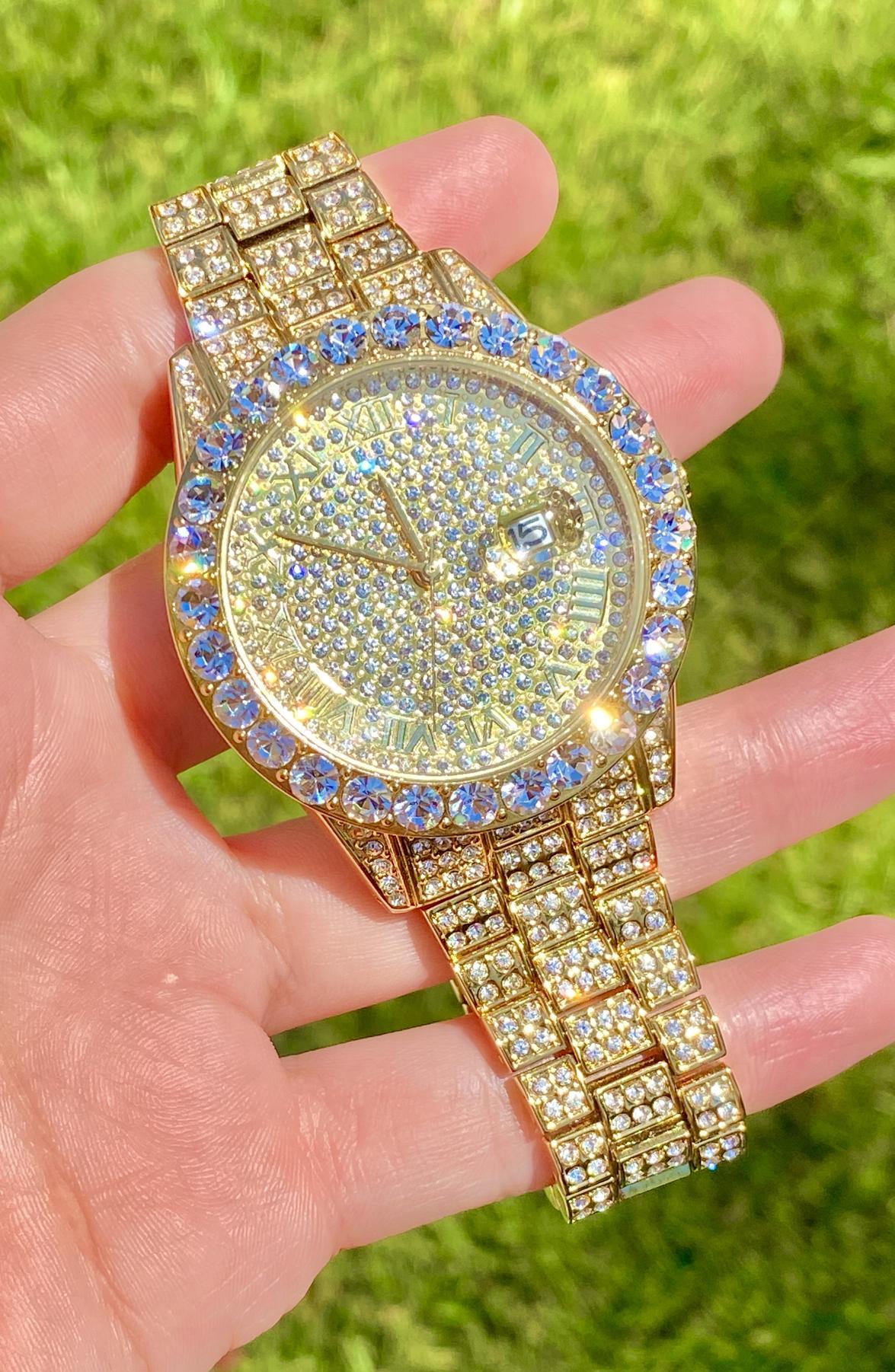 Gold-tone women's and men's watch with a diamond-studded bezel and a sparkling crystal-encrusted band. The watch features a textured gold dial, Roman numeral markers, and a date display at the 3 o'clock position, offering a luxurious and elegant look.