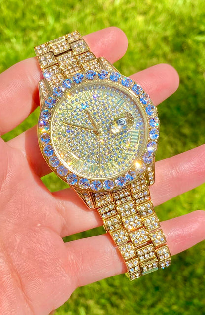 Gold-tone women's and men's watch with a diamond-studded bezel and a sparkling crystal-encrusted band. The watch features a textured gold dial, Roman numeral markers, and a date display at the 3 o'clock position, offering a luxurious and elegant look.