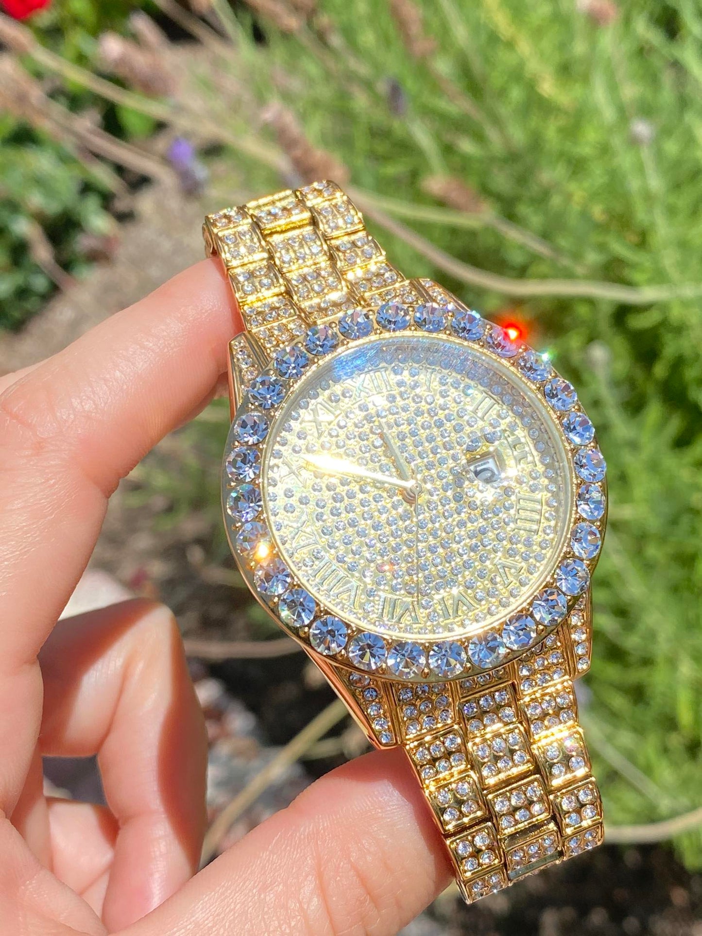 Gold-tone women's and men's watch with a diamond-studded bezel and a sparkling crystal-encrusted band. The watch features a textured gold dial, Roman numeral markers, and a date display at the 3 o'clock position, offering a luxurious and elegant look.