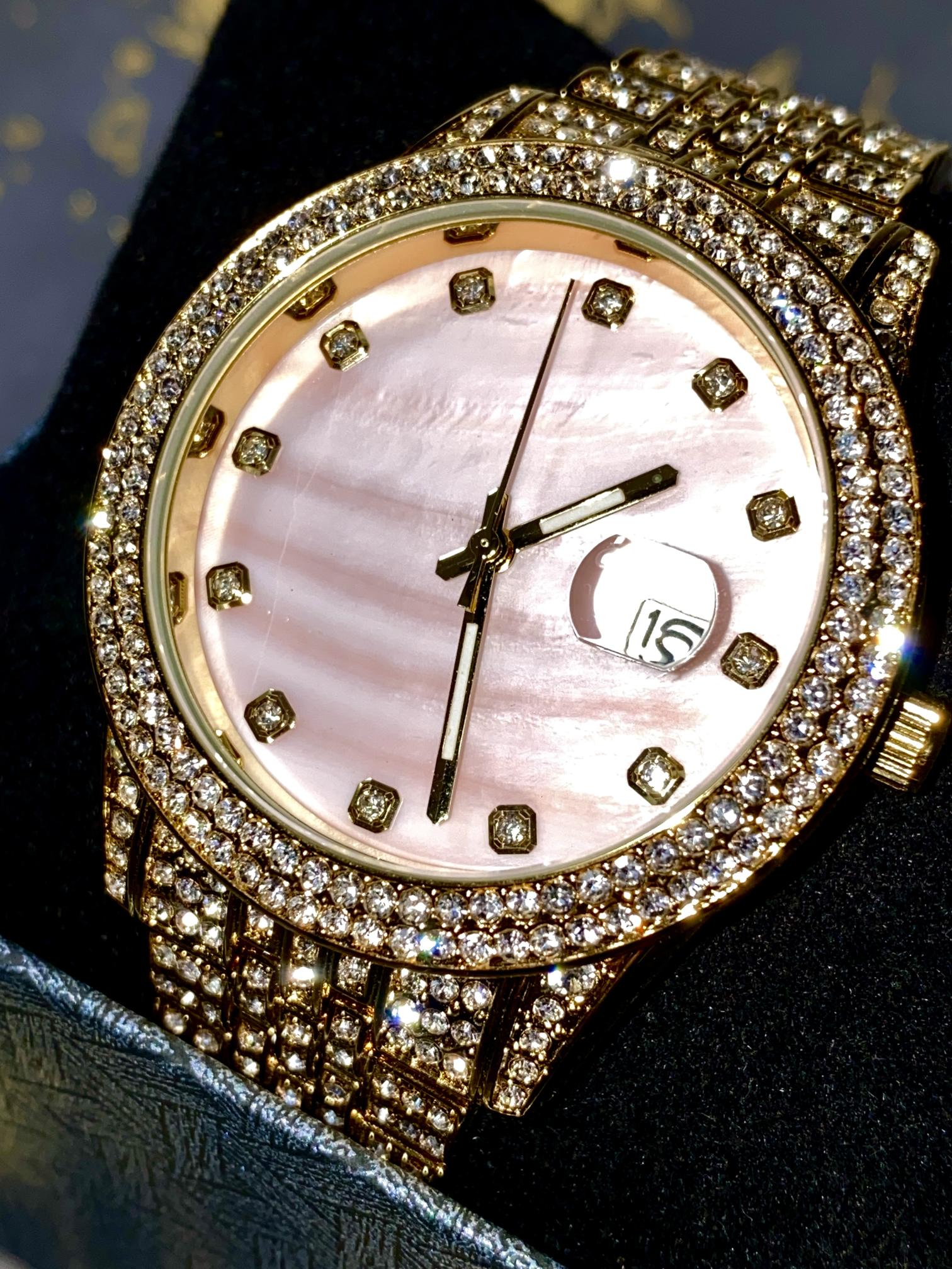 Gold-tone women's watch with a diamond-studded bezel, pink mother-of-pearl dial, and date display at the 3 o'clock position. The watch features a shimmering, crystal-encrusted gold band for a glamorous and luxurious look.